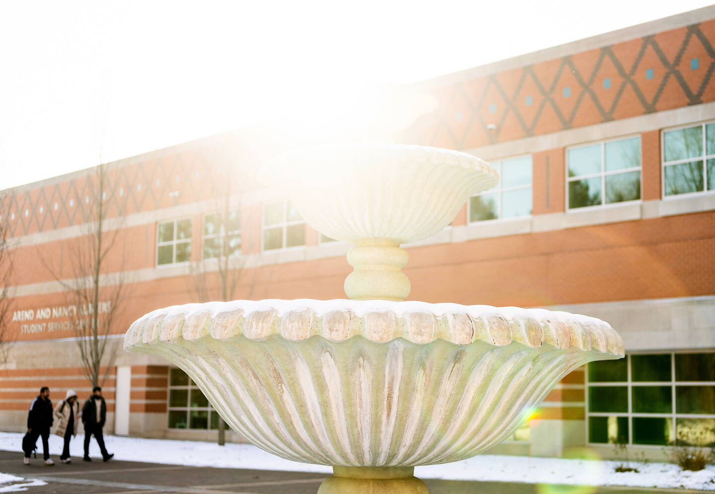 A snow-dusted fountain stands in front of a brick building with people walking by on a sunny winter day.
